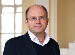 Bild von Sebastian Hess. Ein Mann mit Brille und Anzug lächelt leicht in die Kamera. Hintergrund ist ein Raum mit großen Fenstern und Säulen.