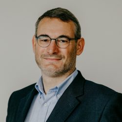 Bild von Rüdiger Wilhelmi. Ein Mann mit Brille und Bart lächelt und trägt ein dunkles Sakko über einem hellblauen Hemd.
