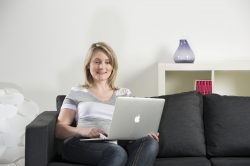 Frau sitzt auf einem Sofa und benutzt einen Laptop. Eine Vase und Bücher sind im Hintergrund auf einem Regal sichtbar.
