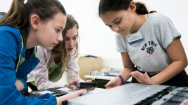 Drei Mädchen arbeiten zusammen an einem Projekt, fokussiert mit technischen Geräten auf einem Tisch.