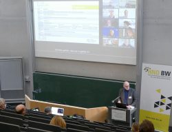 Ein Vortragender spricht vor Publikum in einem Hörsaal, während eine Präsentation mit Programmpunkten projiziert wird.