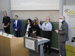Eine Gruppe von sechs Personen steht in einem Raum vor einem Podium, eine Person spricht ins Mikrofon.