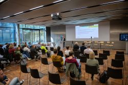Ein Vortrag in einem modernen Konferenzraum. Menschen sitzen auf Stühlen vor einem Referenten und einer Präsentation.