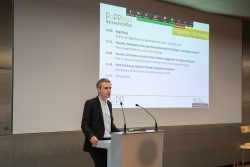 Ein Mann hält einen Vortrag beim PePP 2023 Netzwerktreffen; Programm wird auf einer Leinwand gezeigt.