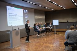 Eine Podiumsdiskussion mit sechs Personen über digitale Prüfungen, ein Mann spricht neben einem Rednerpult, Präsentation projiziert.