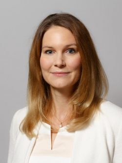 Bild Silke Hertel. Lächelnde Frau mit langen, blonden Haaren in einem weißen Blazer vor grauem Hintergrund.