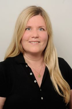 Bild von Susanne Falk. Eine Frau mit langem blondem Haar lächelt. Sie trägt ein schwarzes Hemd und eine Halskette mit Anhänger.