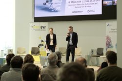 Zwei Personen stehen auf einer Bühne, ein Mann spricht ins Mikrofon. Publikum sitzt davor. Präsentation auf Bildschirm im Hintergrund.