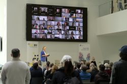 Eine Frau spricht zu einem Publikum in einem Raum, während auf einem Bildschirm viele Personen in einem Videocall zu sehen sind.