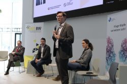 Eine Gruppe von vier Personen auf einer Konferenzbühne; eine Person spricht ins Mikrofon, andere sitzen.
