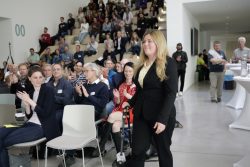 Eine Frau im schwarzen Anzug läuft durch einen Saal, während ein Publikum klatscht. Im Hintergrund sind Menschen auf Treppen.