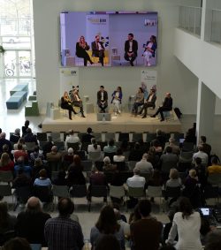 Eine Podiumsdiskussion mit sieben Personen auf der Bühne, vor einem Publikum in einem modernen Konferenzraum.