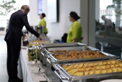Eine Person bedient sich an einem Buffet mit Backwaren. Im Hintergrund stehen zwei Personen an einem Tisch.