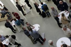 Menschen unterhalten sich bei Stehtischen in einem hellen Raum, einige stehen, andere sitzen auf Bänken.