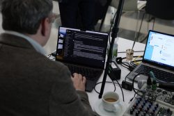 Eine Person arbeitet an einem Laptop, umgeben von Audiogeräten und einer Tasse Kaffee. Ein zweiter Laptop steht daneben.