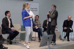 Eine Gruppe von Menschen diskutiert auf einer Bühne. Eine Frau im blauen Blazer spricht mit einem Mann, der ein Mikrofon hält.