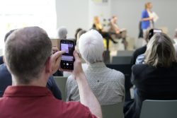 Ein Mann im Publikum fotografiert mit seinem Smartphone eine Präsentation auf einer Bühne bei einem Event.