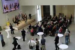Personen in einem Veranstaltungsraum hören einer Podiumsdiskussion zu. Auf der Bühne und auf Bildschirmen sind Redner*innen zu sehen.