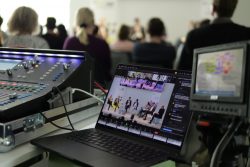 Laptop zeigt eine Konferenz mit mehreren Personen auf einem Podium; daneben Mischpult und Monitor im Vordergrund.