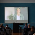 Eine Gruppe von Menschen sitzt in einem Raum und schaut eine Projektion mit einer Frau in einer Videokonferenz an.