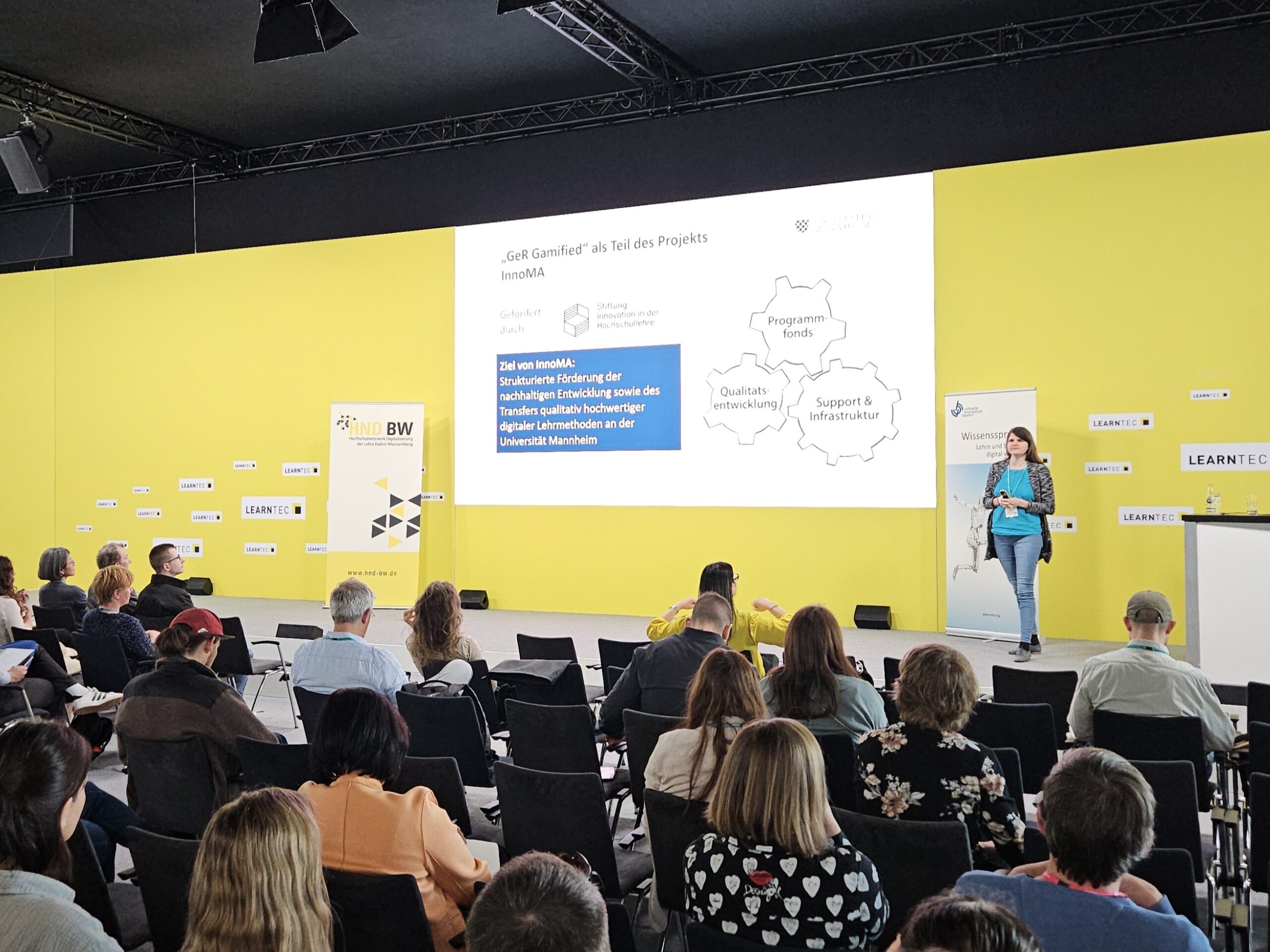 Vortragende auf einer Konferenz präsentiert vor Publikum, vor gelber Wand mit Text und Diagrammen auf großer Leinwand.