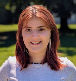 Bild von Emily Möbs. Eine lächelnde Person mit roten Haaren und grauem T-Shirt im Freien bei sonnigem Wetter. Hintergrund unscharf.