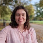 Gergana_Ruseva_2025_04_07 Bild von Gergana Ruseva: Eine Person mit gestreiftem Hemd sitzt lächelnd auf einer Bank im Park mit Bäumen im Hintergrund.
