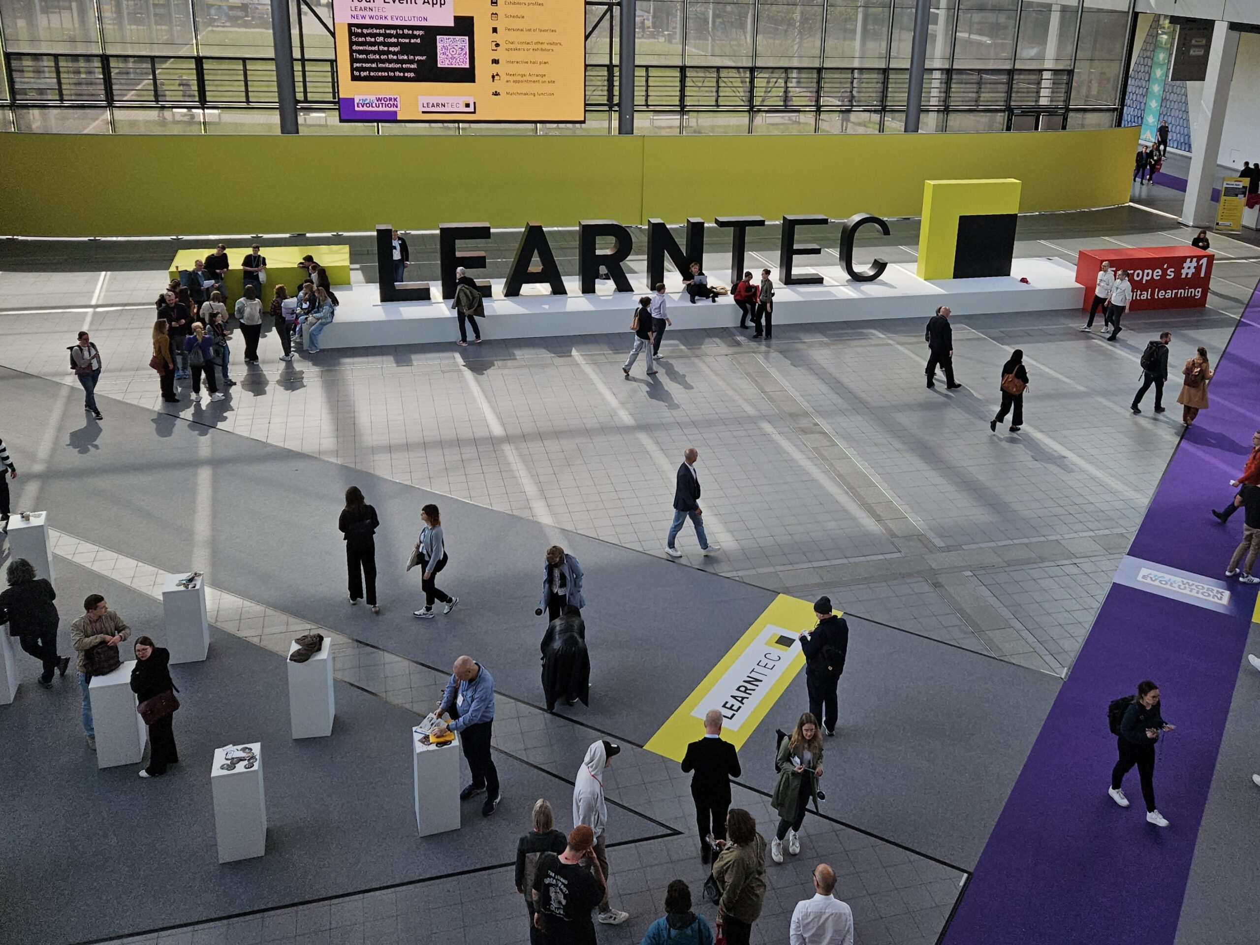 Personen bewegen sich auf einer Messe mit dem Schriftzug LEARNTEC im Hintergrund. Bunte Stände und Plakate sind sichtbar.