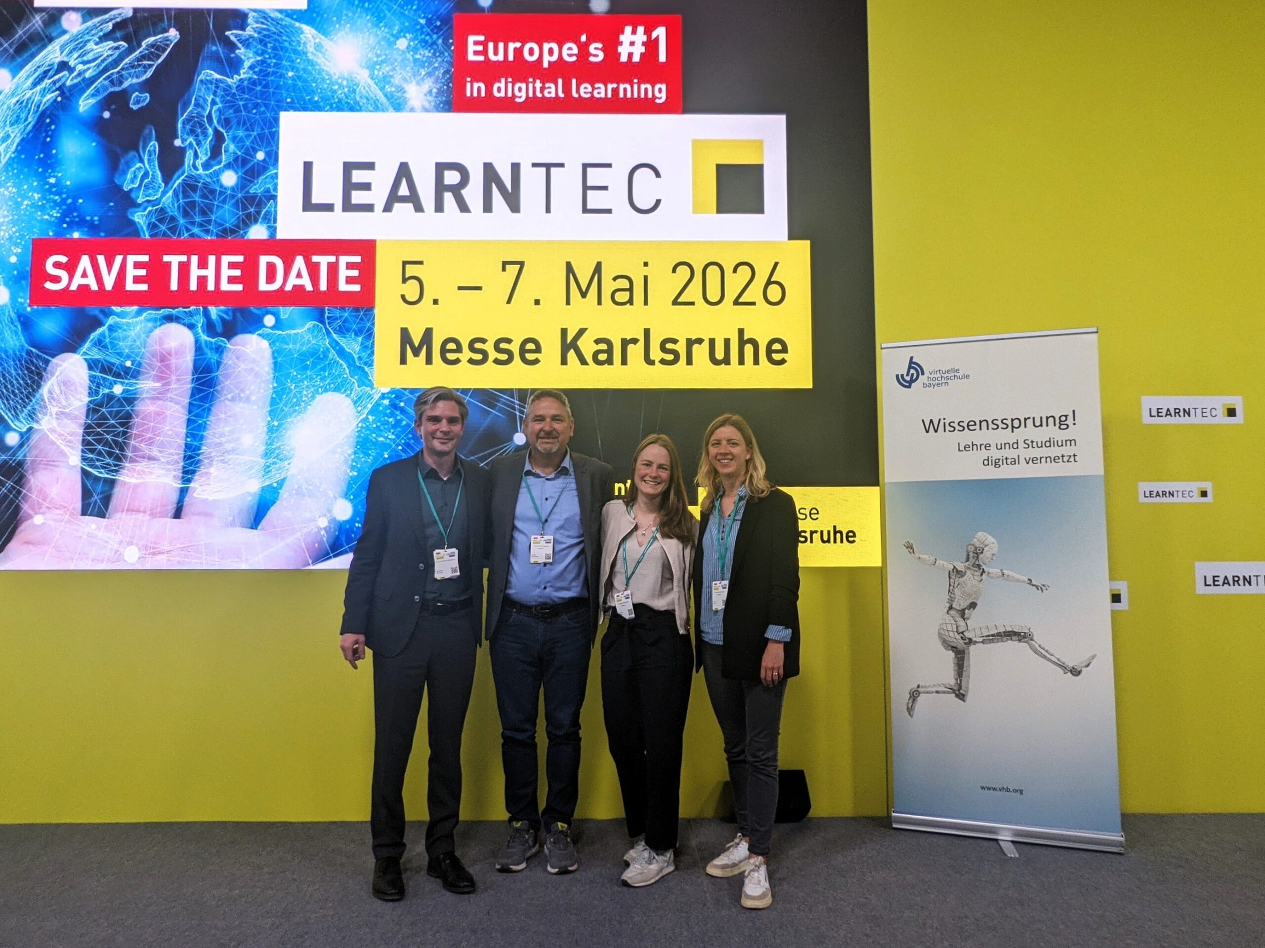 Vier Personen posieren vor einem Messeplakat der LEARNTEC 2026 in Karlsruhe.