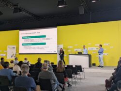 Personen präsentieren auf einer Konferenz vor gelbem Hintergrund über das Umsetzungskonzept bwGPT.