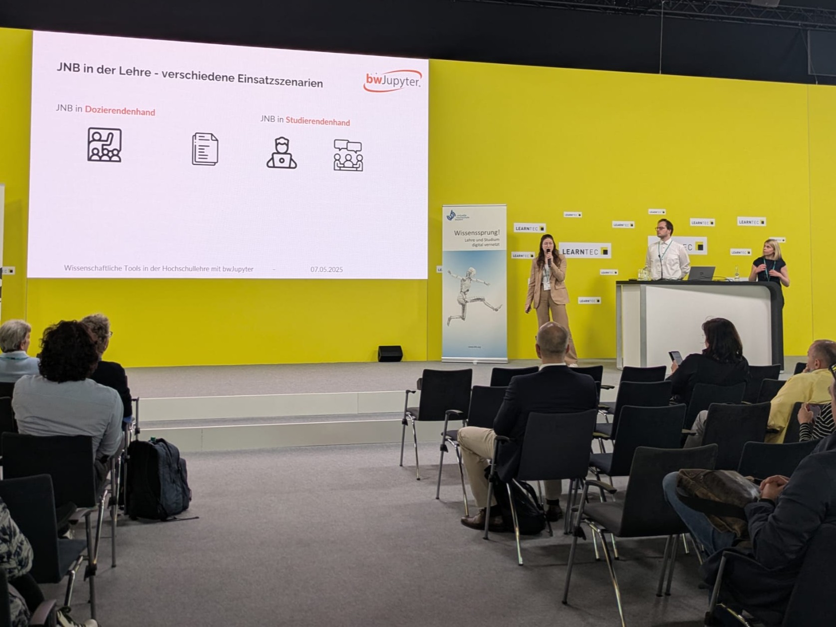 Präsentation über JNB in der Lehre mit Vortragender auf einer Bühne vor einer gelben Leinwand mit Publikum.