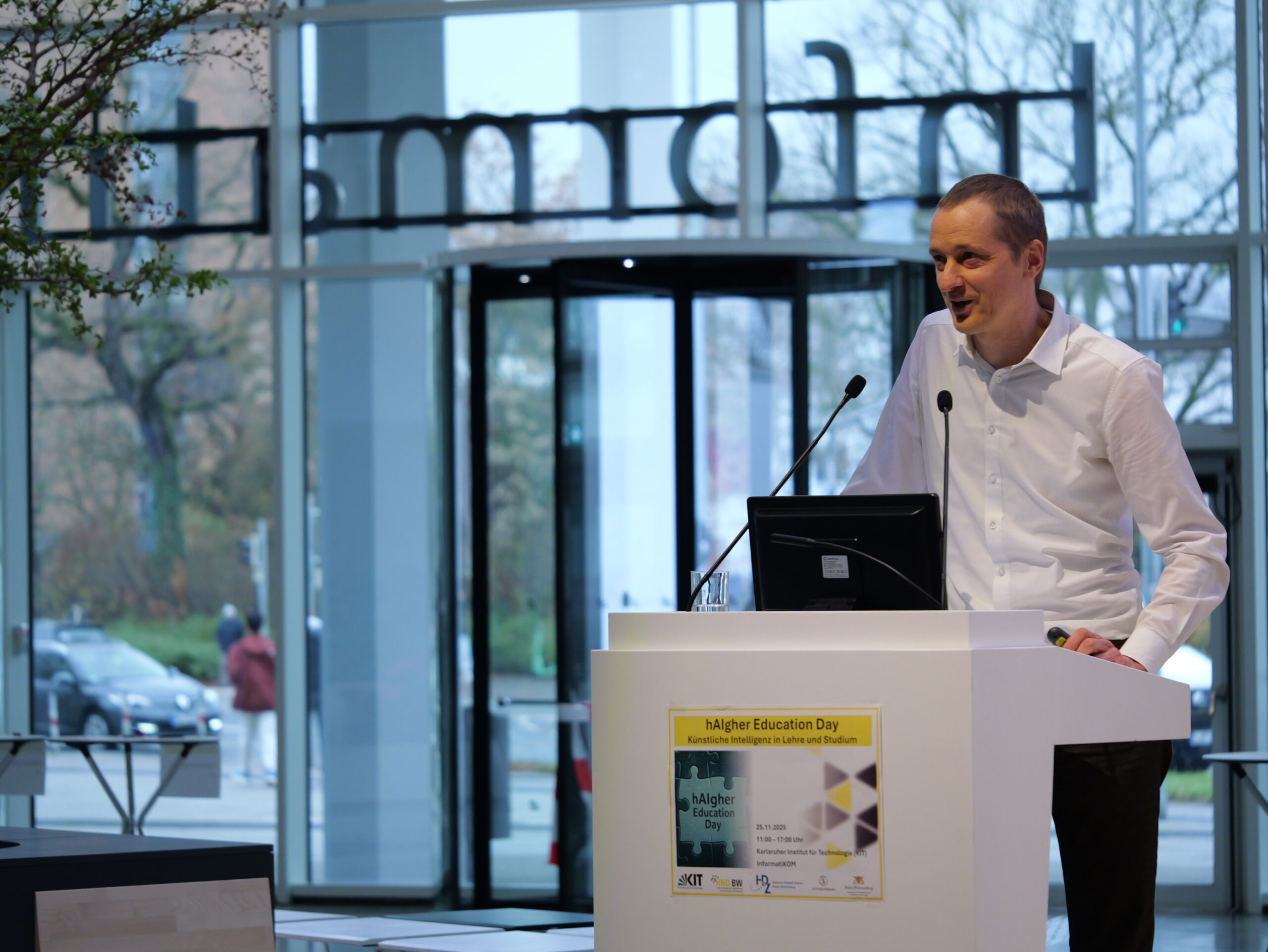 P1002138 - Kopie Ein Mann spricht bei einer Konferenz am Rednerpult. Im Hintergrund ist ein Eingangsbereich mit Glaswänden sichtbar.