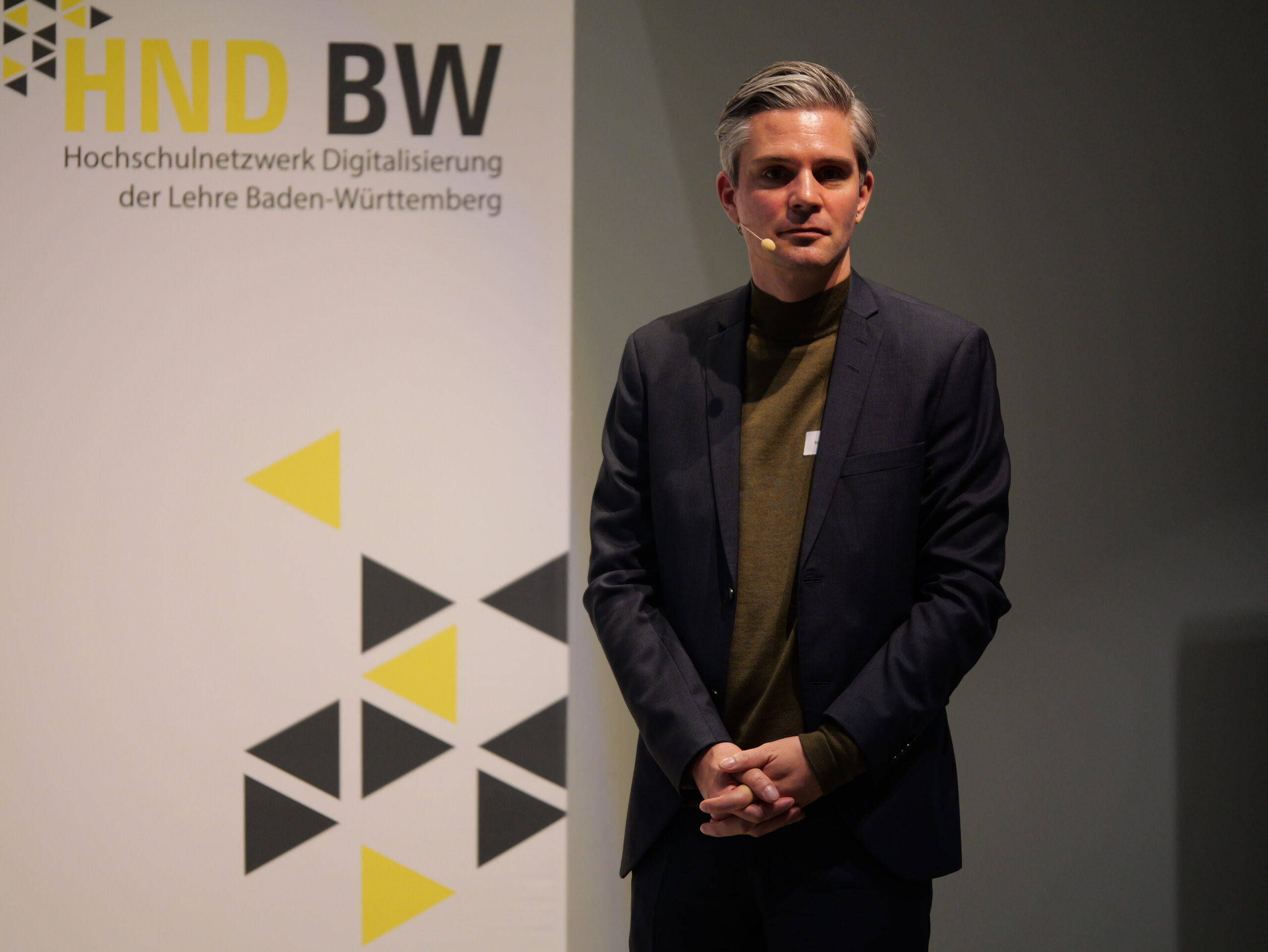 P1002156 - Kopie Eine Person steht vor einem Plakat mit der Aufschrift HND BW Hochschulnetzwerk Digitalisierung der Lehre Baden-Württemberg.