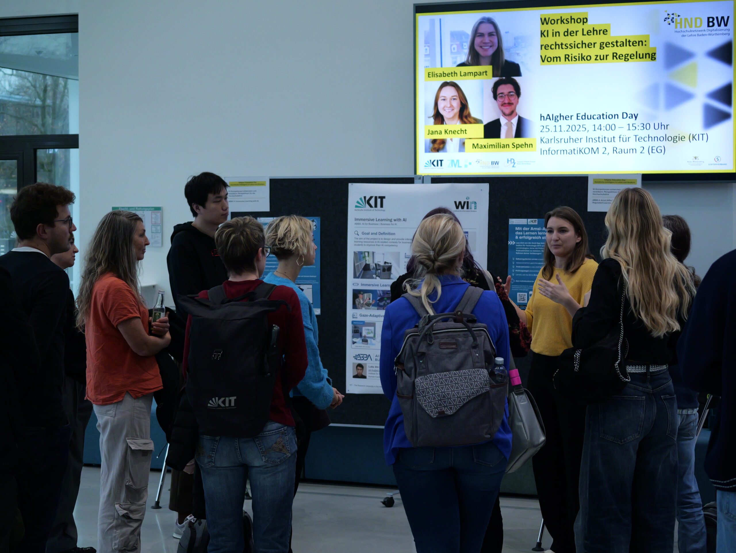 P1002248 - Kopie Gruppe von Menschen diskutiert vor Informationsplakaten in einem Gebäude. Ein Bildschirm kündigt einen Workshop an.