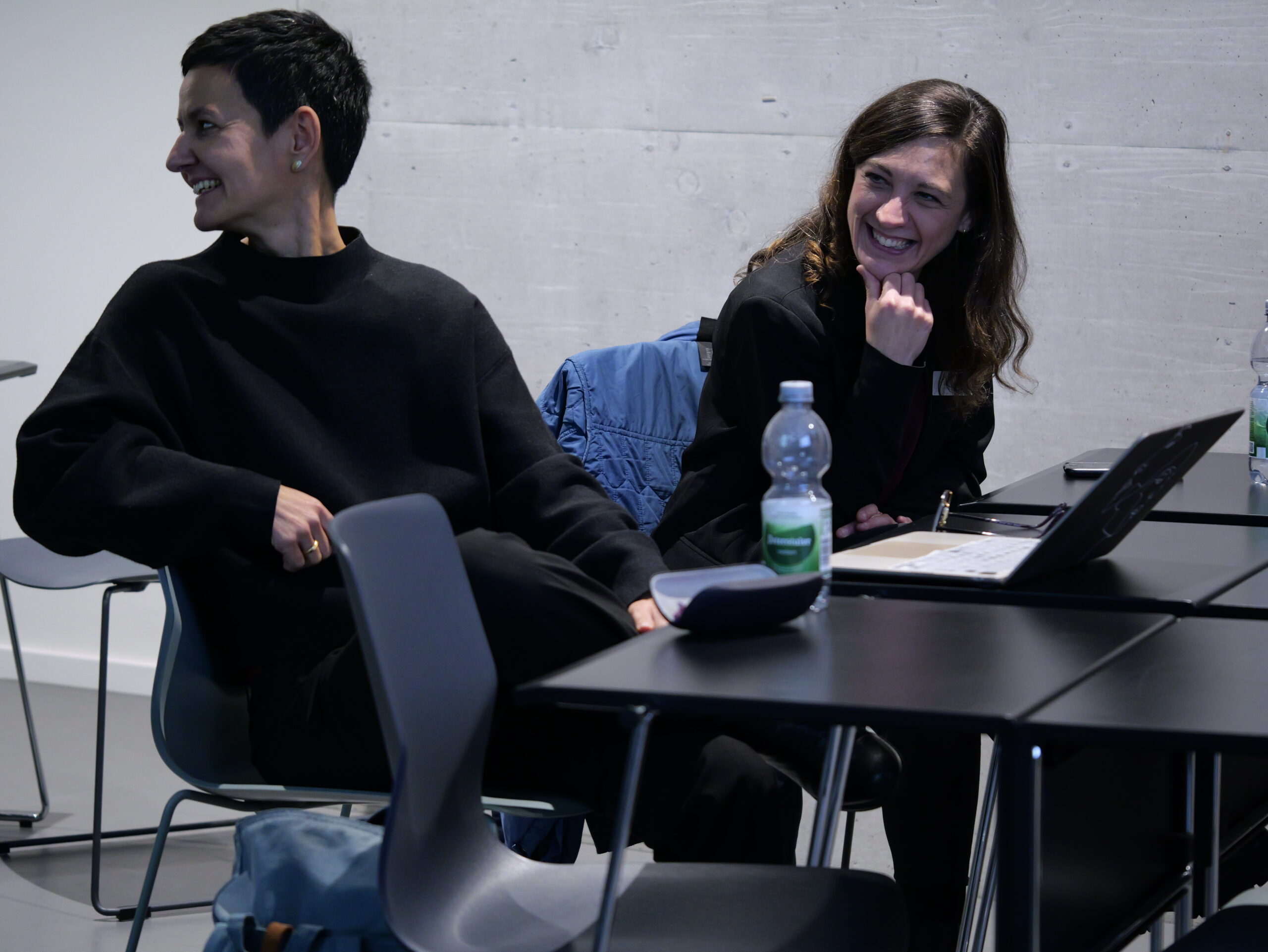 P1002294 Zwei Personen sitzen an einem Tisch und lachen. Vor ihnen stehen Wasserflaschen und ein Laptop auf dem Tisch.