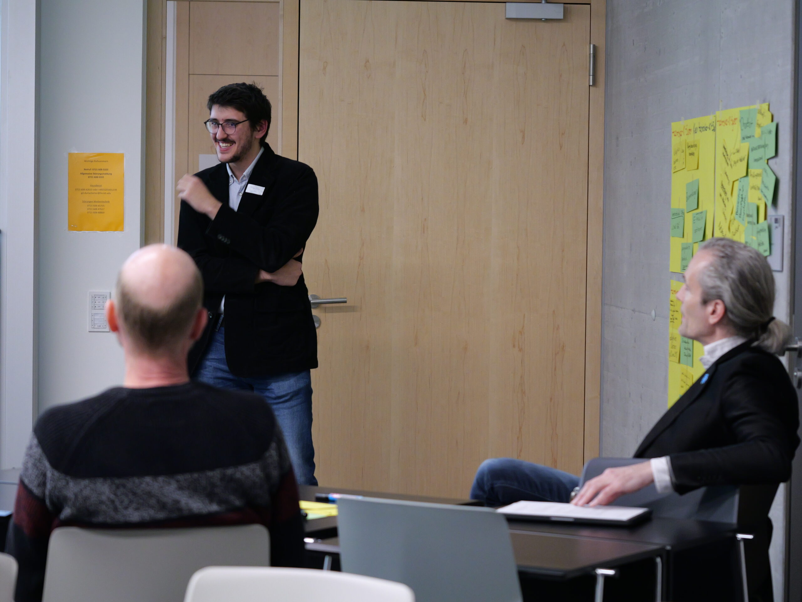 P1002307 Ein Mann steht lächelnd vor einem Raum und spricht zu zwei sitzenden Personen. Notizen sind an der Wand zu sehen.