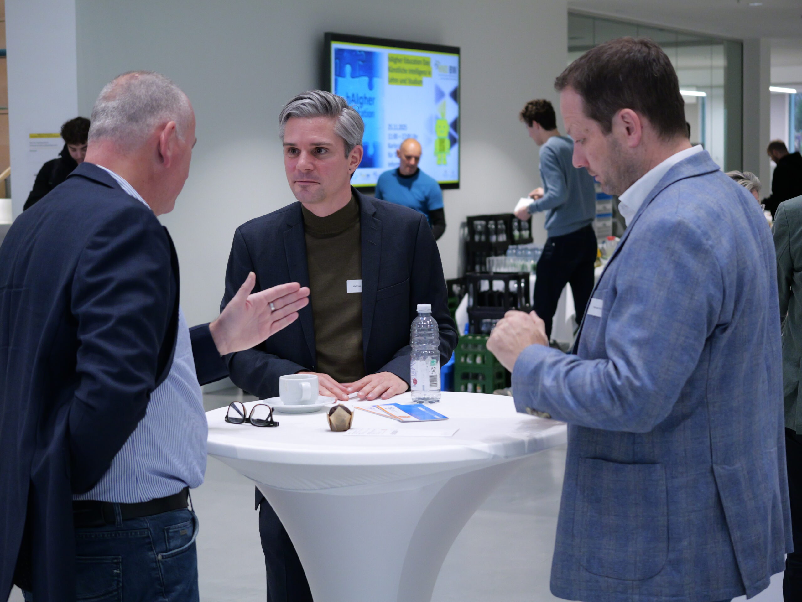 P1002399 Drei Männer im Gespräch an einem Stehtisch in einem modernen Konferenzraum, mit Getränken und Infomaterial auf dem Tisch.