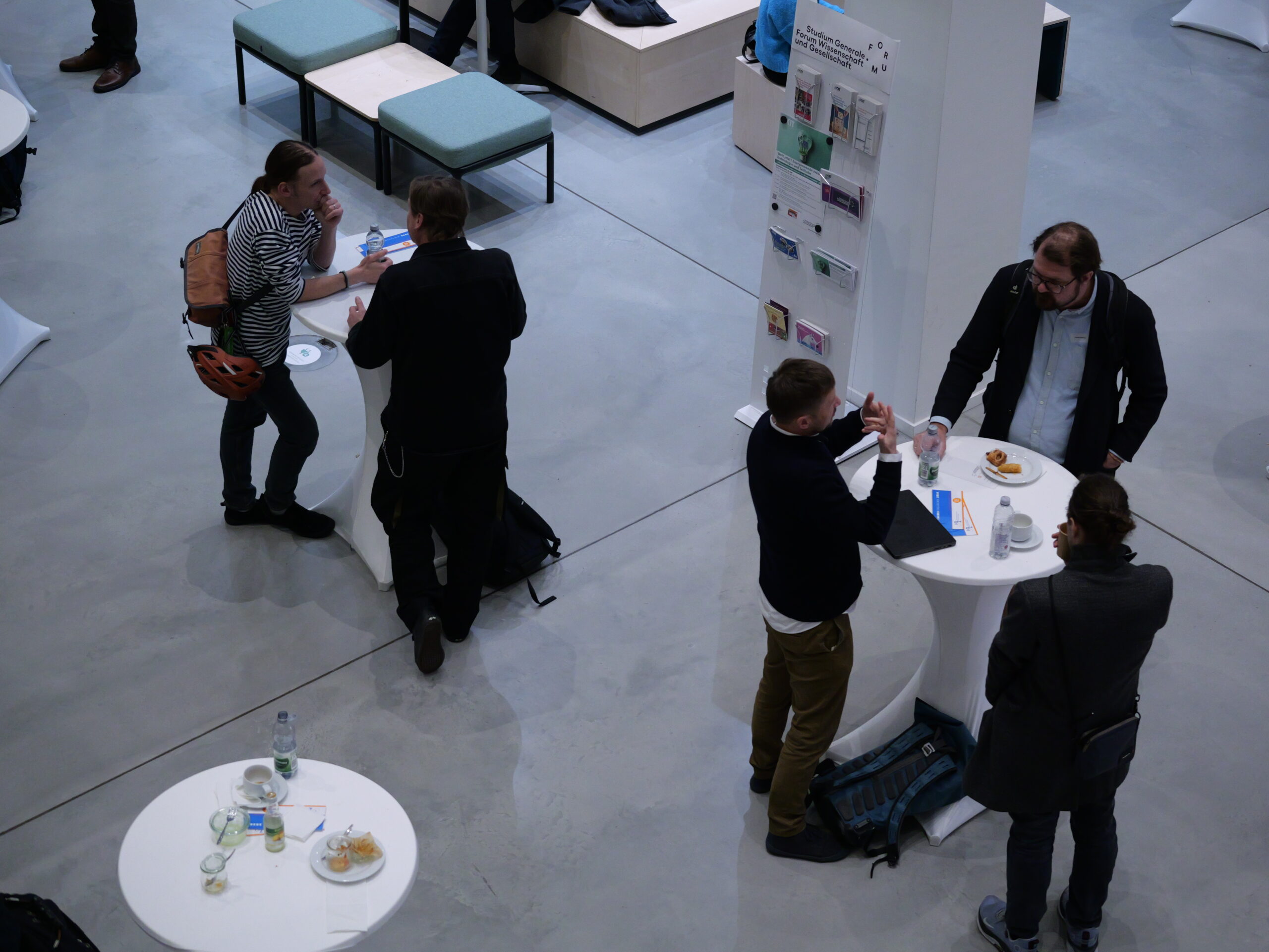 P1002428 - Kopie Menschen stehen an Stehtischen in einem modernen Raum und unterhalten sich, während sie Getränke und Snacks genießen.