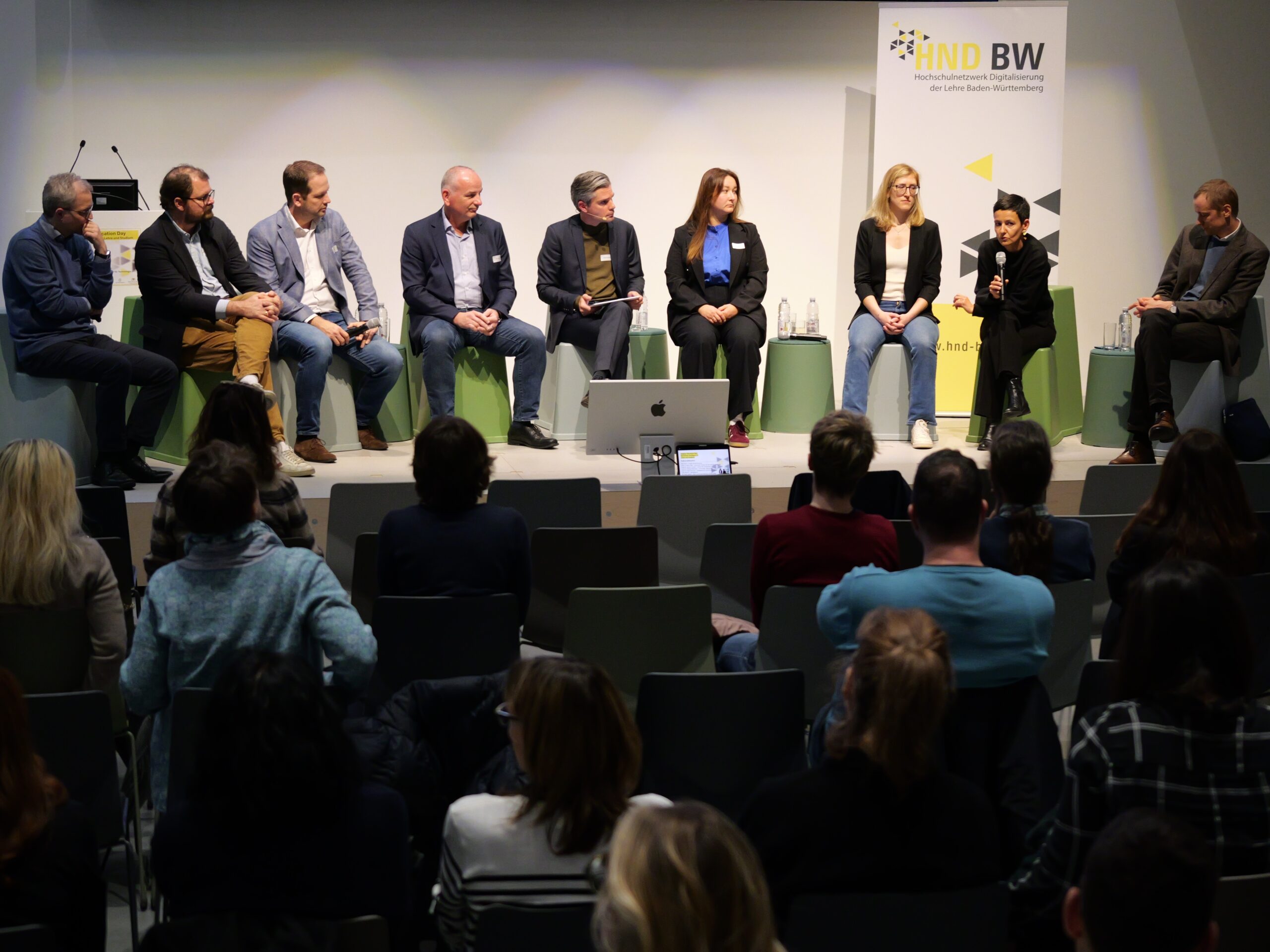 P1002474 - Kopie Eine Gruppe von neun Personen diskutiert auf einer Podiumsdiskussion vor einem Publikum in einem Konferenzraum.