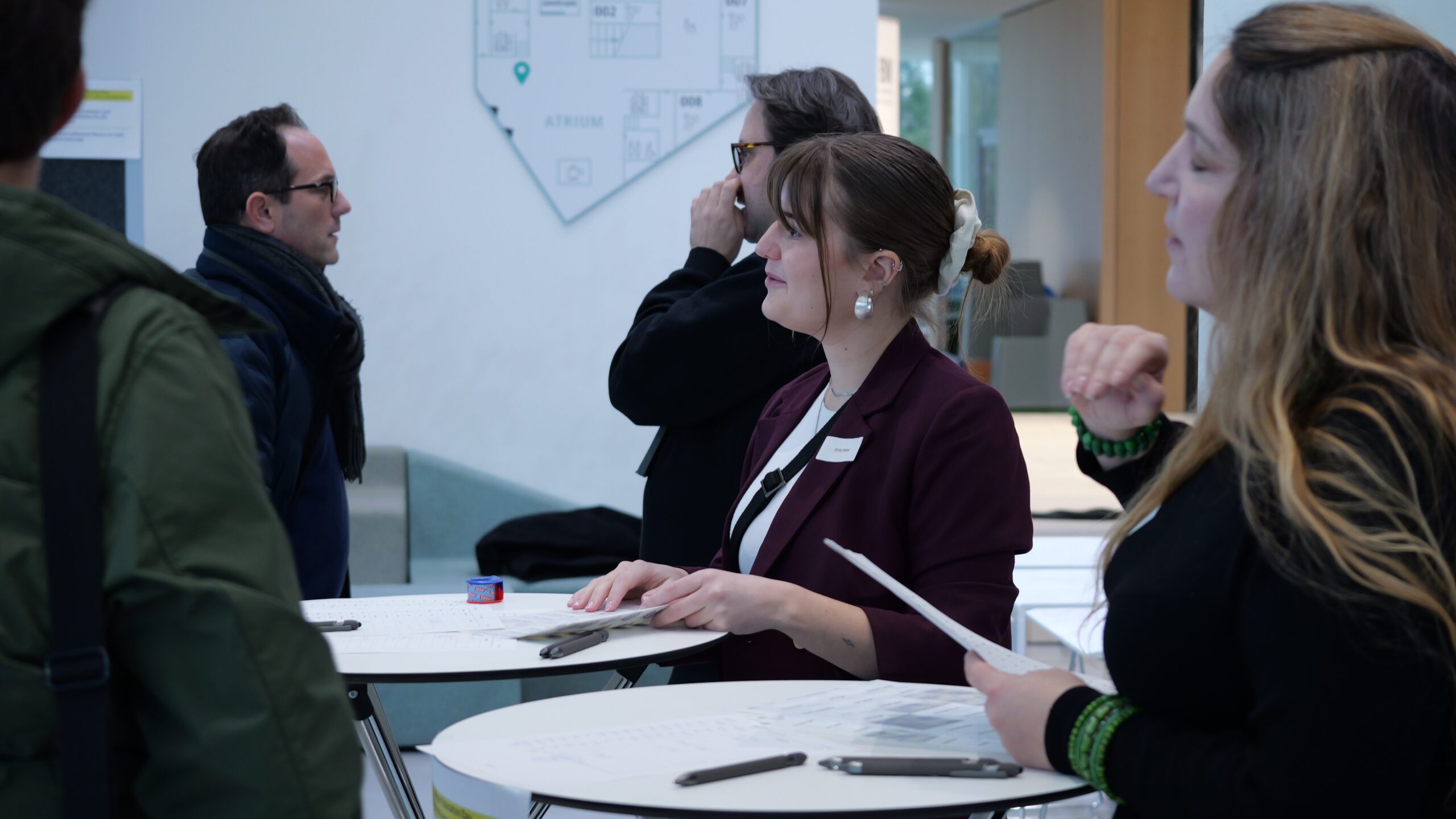 Personen im Gespräch an Stehtischen in einer modernen Lobby, eine Frau schreibt etwas.