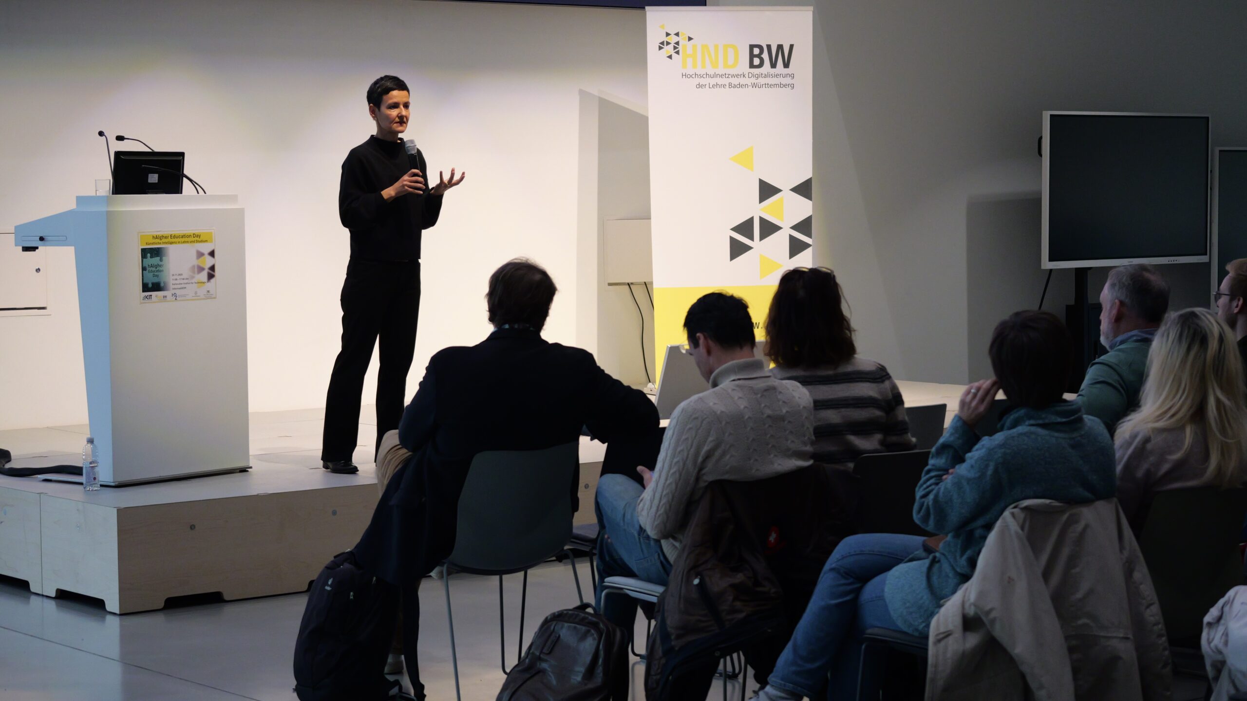 Rednerin hält Vortrag auf einer Bühne vor Zuhörern, neben einem Banner mit der Aufschrift „Hochschulnetzwerk Digitalisierung“.