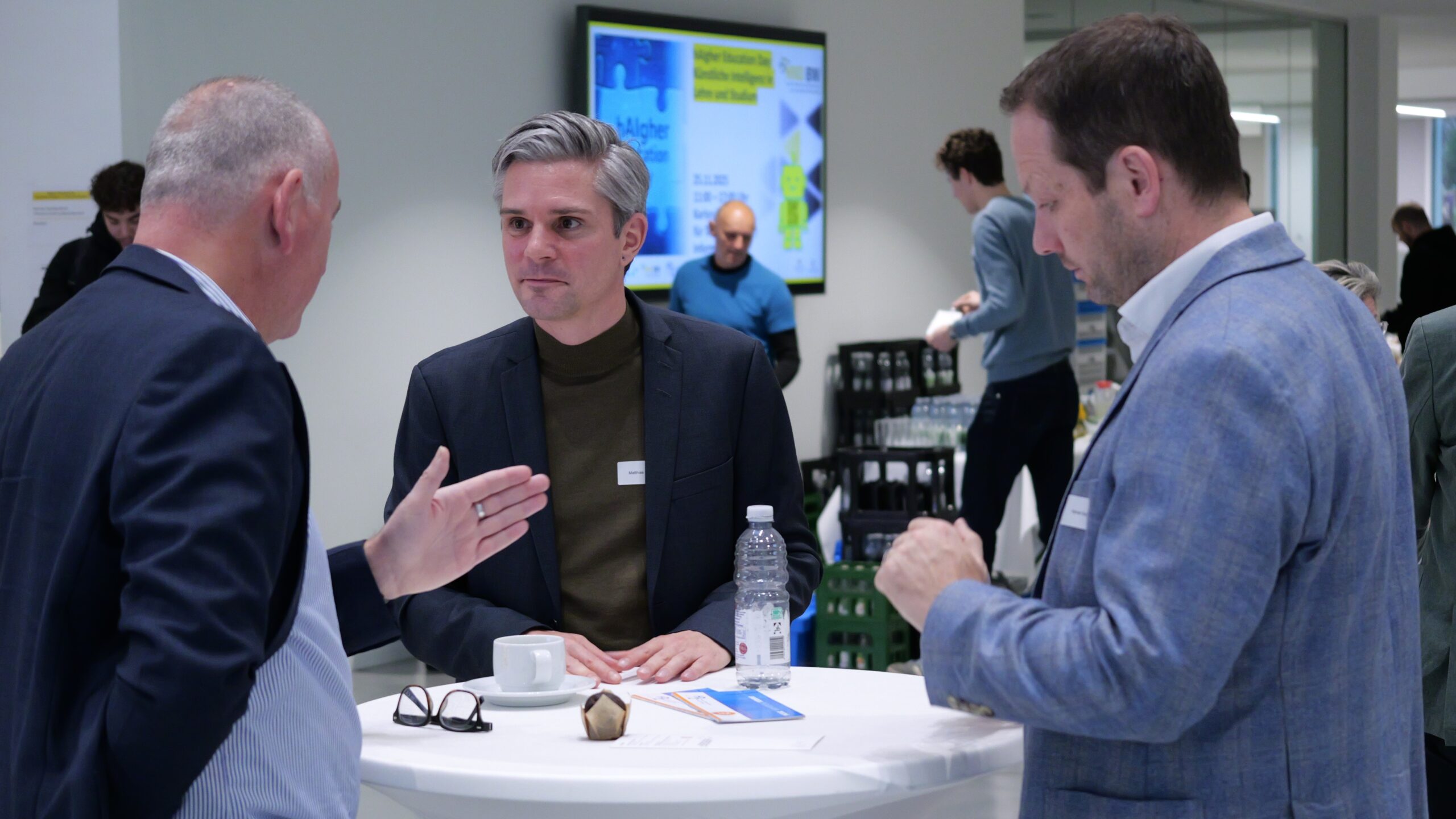 Drei Männer sprechen stehend an einem Tisch bei einem Networking-Event. Im Hintergrund stehen Getränke bereit.