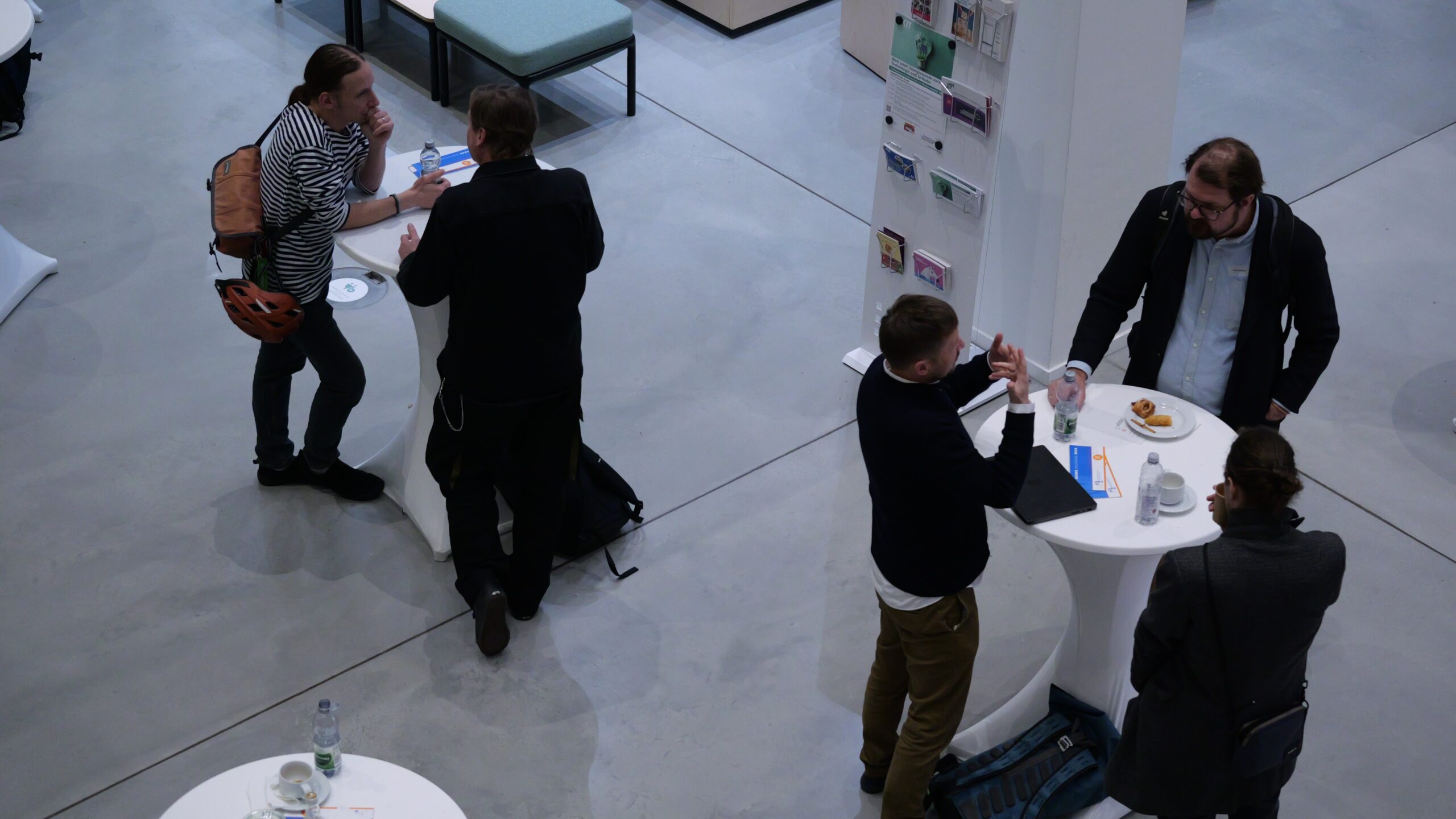 Vier Personen unterhalten sich in einer modernen Lobby an Stehtischen, einige mit Getränken und Snacks.