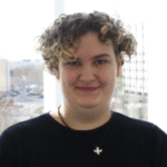 Eine Person mit lockigem Haar lächelt in die Kamera. Im Hintergrund sind unscharfe Gebäude und Bäume zu sehen.