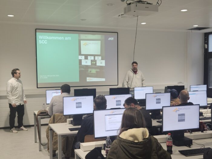 Schulung im Computerraum. Zwei Personen präsentieren vor Teilnehmern, die an Computern sitzen. Bildschirm zeigt Willkommen