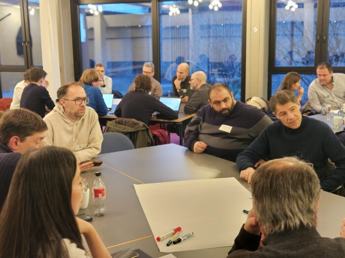Eine Gruppe von Menschen sitzt bei einer Besprechung an Tischen mit Laptops und Notizen in einem Konferenzraum.