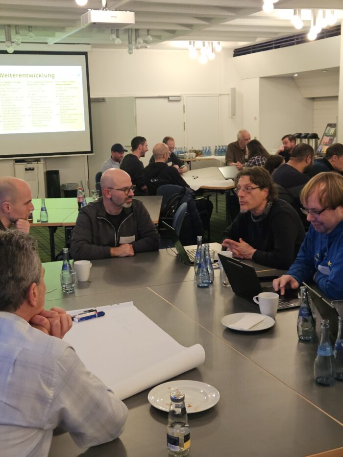 Eine Gruppe von Menschen in einem Meetingraum diskutiert um einen Tisch mit Laptops und Getränken. Präsentation im